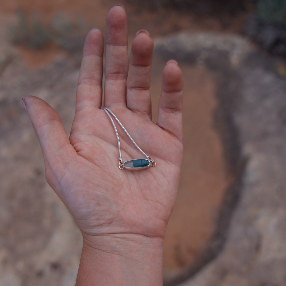 Hand holding a silver necklace with a blue pendant against a natural background